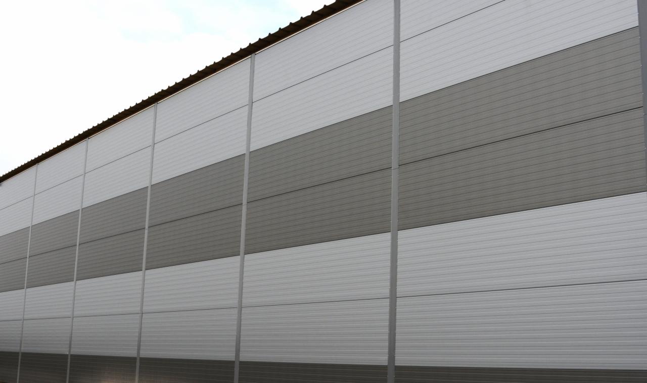 Szara ściana hali przemysłowej wykonana z paneli warstwowych ułożonych w poziome pasy o naprzemiennych odcieniach szarości, widoczna z dołu pod kątem.