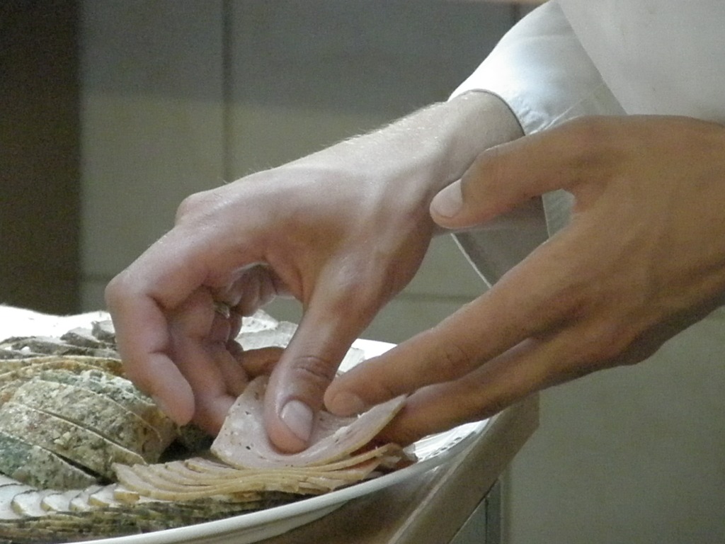 Kucharz układa plasterki wędliny na talerzu, obok pokrojona pieczeń z ziołami.