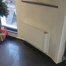 Biały grzejnik panelowy zamontowany pod oknem obok małej choinki, widok z boku, czarna podłoga z płytek