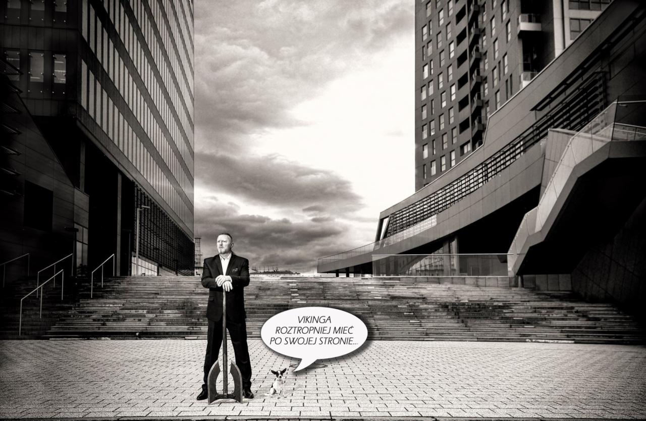 Czarno-białe zdjęcie mężczyzny w garniturze opartego o stylizowany trójząb, z psem u stóp, na tle nowoczesnej architektury biurowej i pochmurnego nieba, z dymkiem dialogowym z tekstem 'Vikinga...