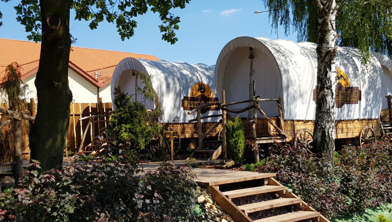 Dwa wozy kempingowe w stylu western, z drewnianymi schodami i roślinnością, ustawione na zewnątrz budynku z pomarańczowym dachem.