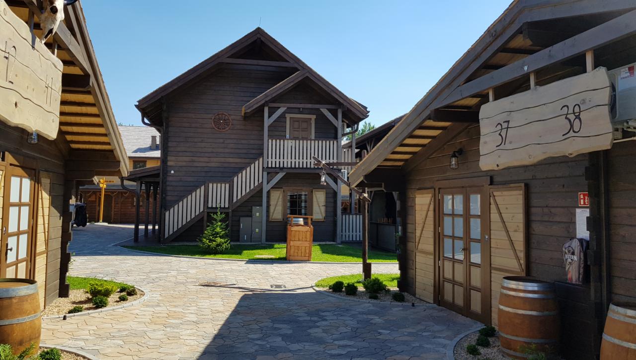 Kompleks budynków w stylu western z drewnianą elewacją, numerami 37 i 38 na tablicach, beczkami przy wejściu i kamienną ścieżką.