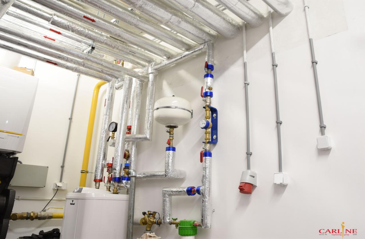 Instalacja grzewcza z kotłem, rurami izolowanymi folią aluminiową, zbiornikiem wyrównawczym i osprzętem, widoczna w pomieszczeniu technicznym z białymi ścianami i żółtą rurą gazową.