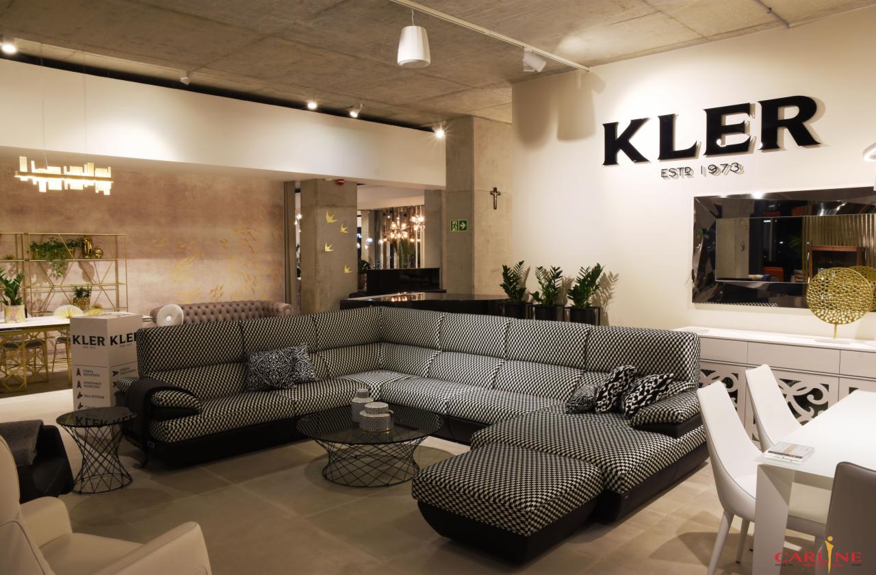 Przestronne wnętrze salonu meblowego z narożną sofą w czarno-białą pepitkę, stolikiem kawowym ze szklanym blatem i białymi krzesłami przy stole, oświetlone nowoczesnymi lampami, z widocznym logo...