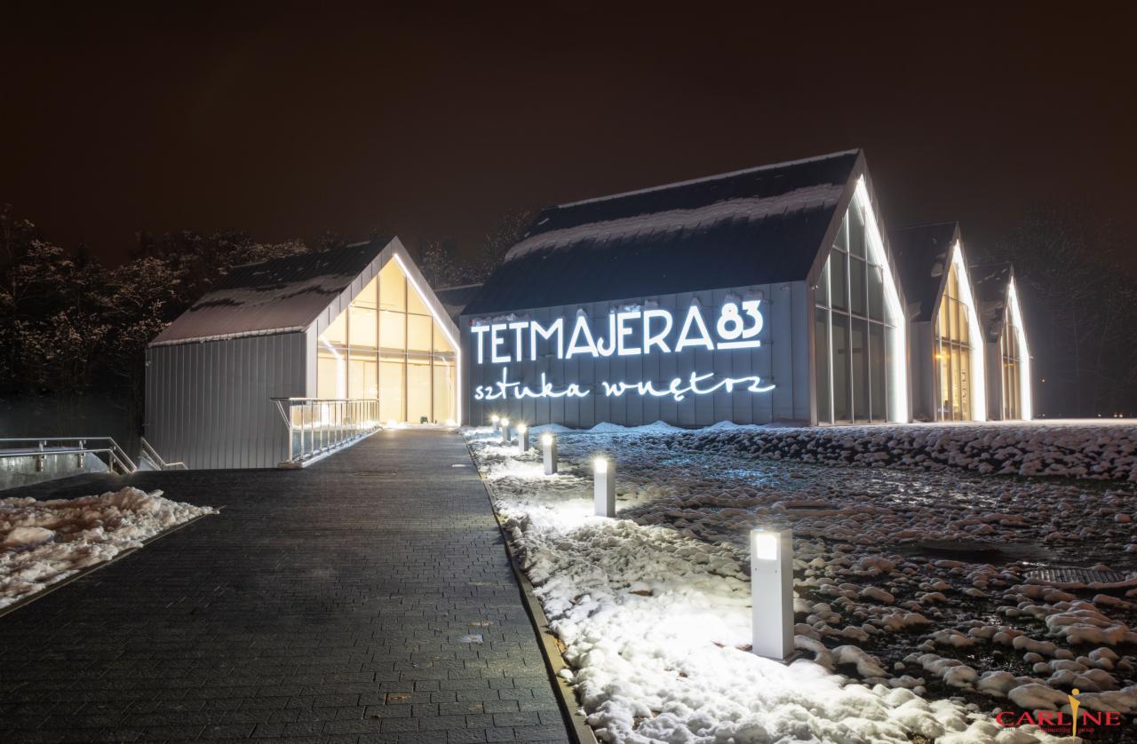 Nowoczesny budynek z przeszklonymi ścianami i oświetlonym logo 'TETMAJERA 83 sztuka wnętrz' w zimowej scenerii, oświetlony chodnik prowadzi do wejścia.