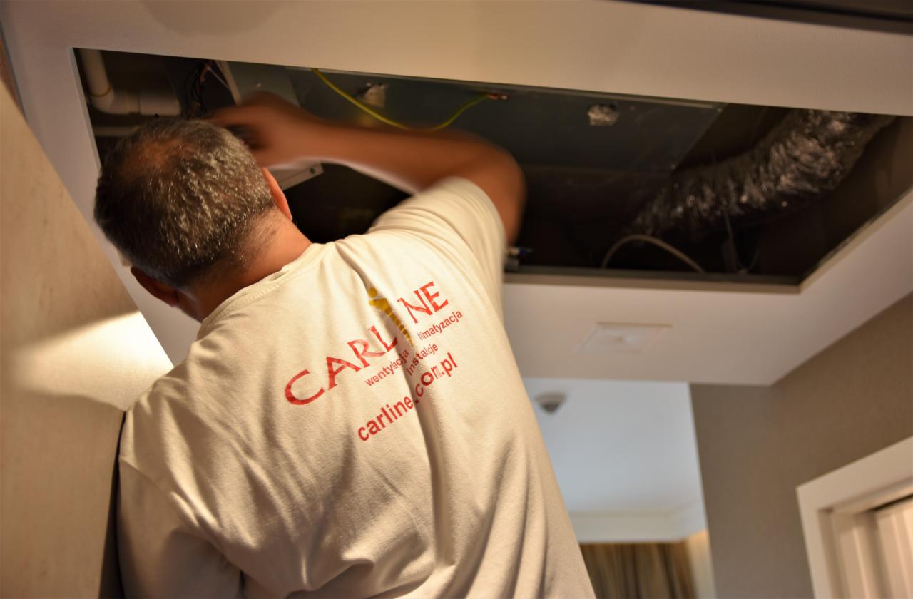 Instalator w białej koszulce z logo firmy, pracujący przy instalacji wentylacyjnej pod sufitem podwieszanym, widoczny przewód wentylacyjny.