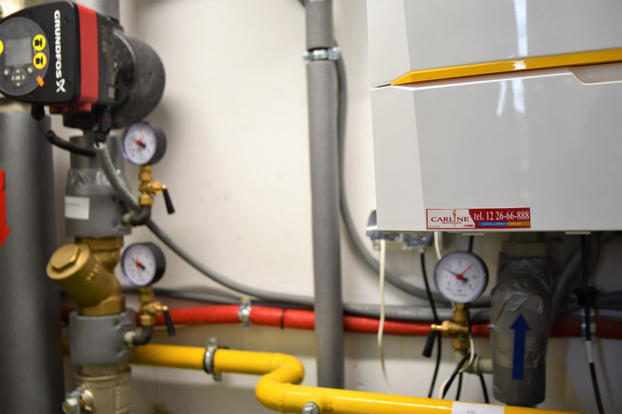Fragment instalacji grzewczej z kotłem, pompą obiegową Grundfos i rurami w kolorach żółtym i czerwonym, widoczne manometry i zawory.