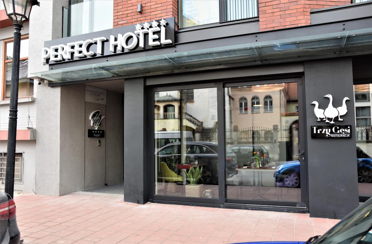 Wejście do hotelu Perfect Hotel i restauracji Trzy Gęsi z widocznymi logotypami i elementami wystroju wnętrz w odbiciu szyby.