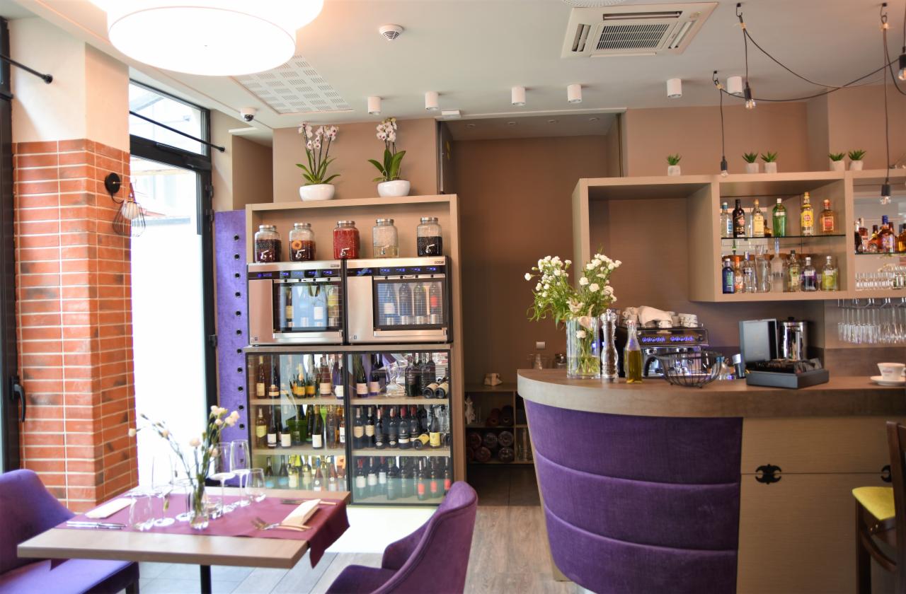 Wnętrze lokalu gastronomicznego z regałami chłodniczymi na wino, nowoczesnym barem z ekspresami do kawy i stolikiem z nakryciem w fioletowej tonacji.