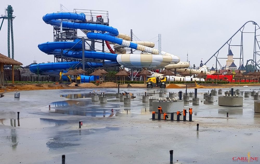 Budowa parku wodnego: widok na betonowe fundamenty pod atrakcje wodne, w tle zjeżdżalnie i roller coaster, mała walcarka do ubijania piasku w tle.