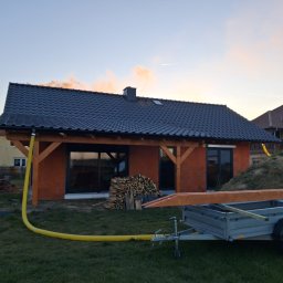 SW-BAU Usługi Budowlane Jolanta Willim - Niski dom w trakcie budowy z czerwonej cegły, z czarnym dachem i drewnianą konstrukcją. Na trawniku przyczepa z drewnem i żółty wąż. Zachodzące słońce.