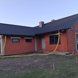 SW-BAU Usługi Budowlane Jolanta Willim - Niedokończony dom z czerwonej cegły z czarnym dachem, widoczne okna i żółte rury wentylacyjne. Częściowo zagospodarowany teren wokół budynku.