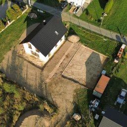 SW-BAU Usługi Budowlane Jolanta Willim - Zdjęcie z drona: biały dom z czarnym dachem w trakcie budowy, widoczne przygotowane fundamenty i teren wokół, w tle zieleń i zabudowa jednorodzinna.