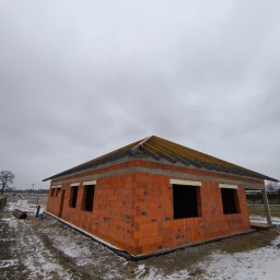 SW-BAU Usługi Budowlane Jolanta Willim - Surowy, ceglany dom w budowie, z widoczną konstrukcją dachu i oknami bez szyb, otoczony zimowym krajobrazem. Ujęcie z niskiej perspektywy podkreśla bryłę budynku.