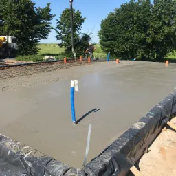 Świeżo wylana, gładka wylewka betonowa na budowie, z widocznym zbrojeniem, rurami instalacyjnymi i miksokretem w tle, zabezpieczona folią.