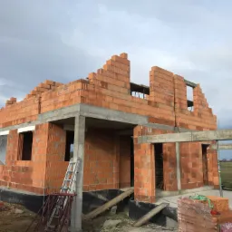 Budowa domu murowanego z czerwonej cegły ceramicznej, widoczne fragmenty szalunków, drabina oparta o ścianę, niebieska beczka i paleta cegieł na placu budowy w pochmurny dzień.
