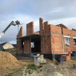 Wznoszenie ścian z czerwonej cegły na budowie domu jednorodzinnego, z widocznym podnośnikiem teleskopowym Bobcat, stertą piasku, taczką i betoniarką.