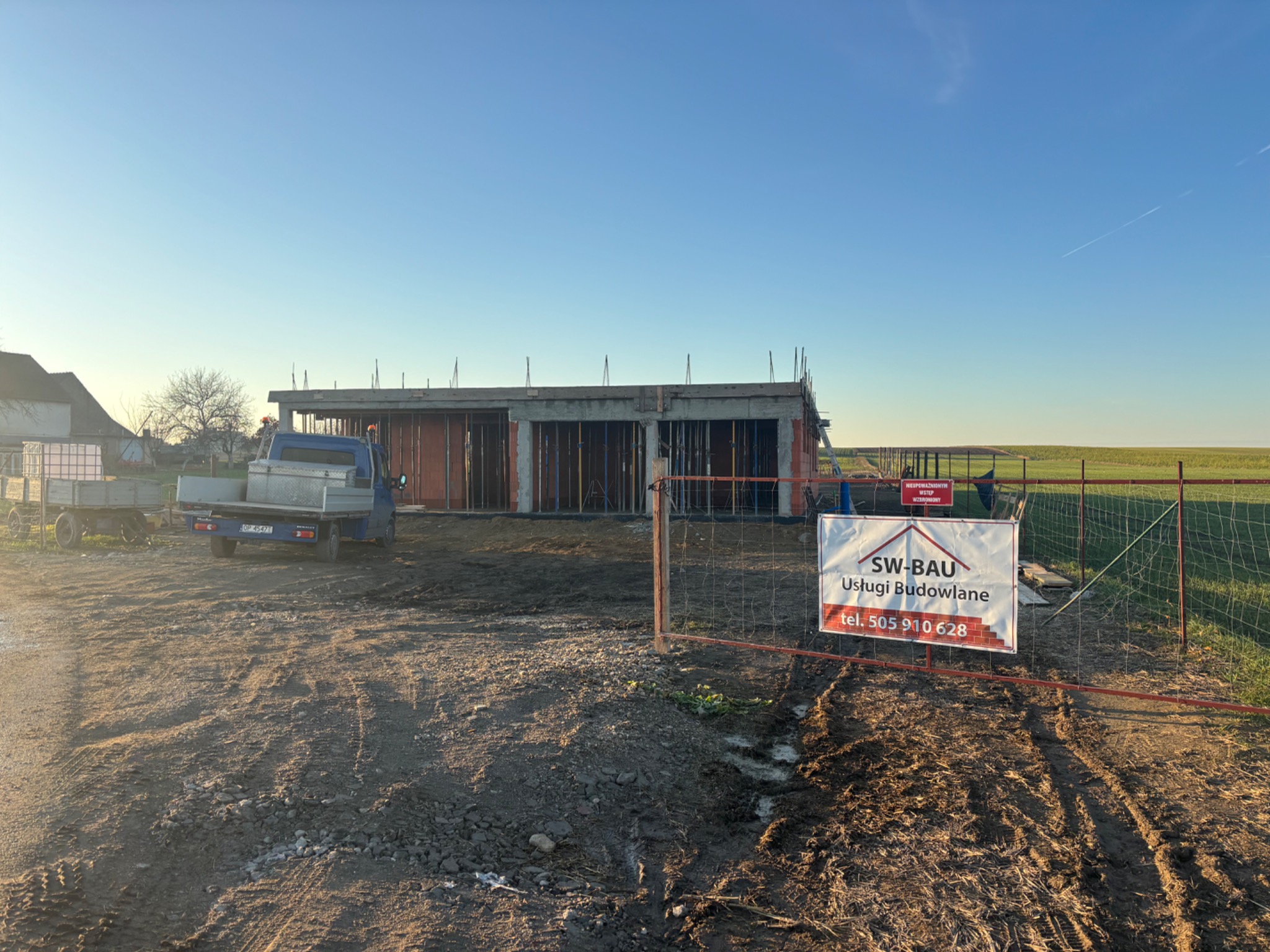 Budowa domu w stanie surowym otwartym, widok z zewnątrz przez ogrodzenie z tablicą informacyjną firmy budowlanej. Na posesji stoi samochód dostawczy.