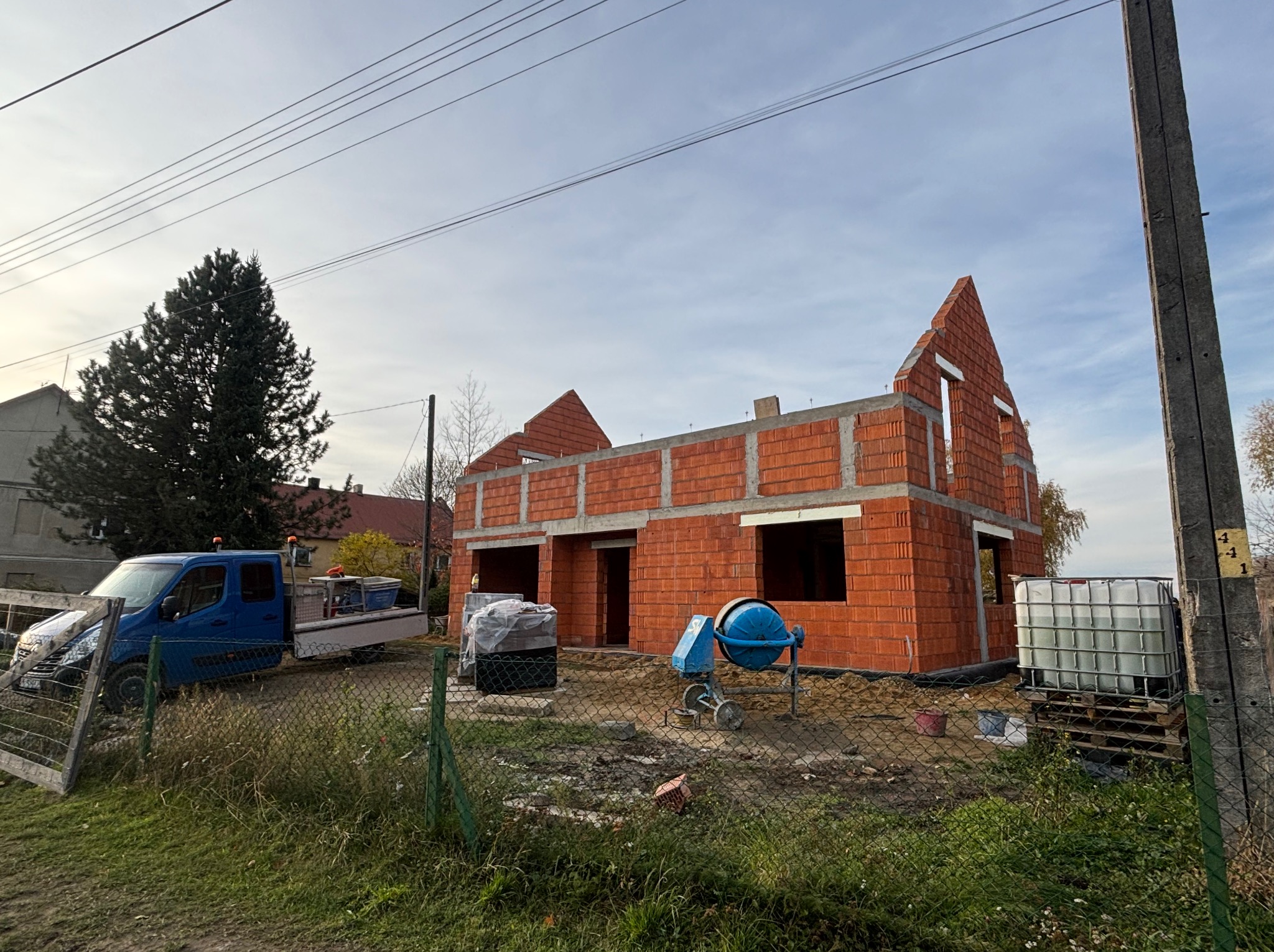 Wznoszenie domu z czerwonej cegły, widoczna betoniarka i auto dostawcze. Budowa w toku, ogrodzenie z siatki, pochmurne niebo w tle.