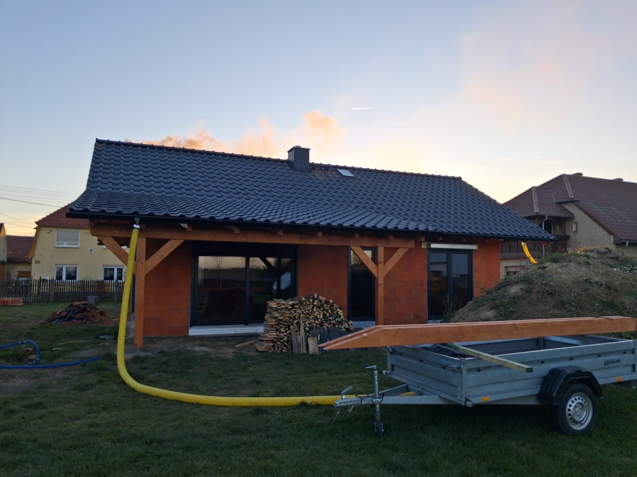Niski dom w trakcie budowy z czerwonej cegły, z czarnym dachem i drewnianą konstrukcją. Na trawniku przyczepa z drewnem i żółty wąż. Zachodzące słońce.