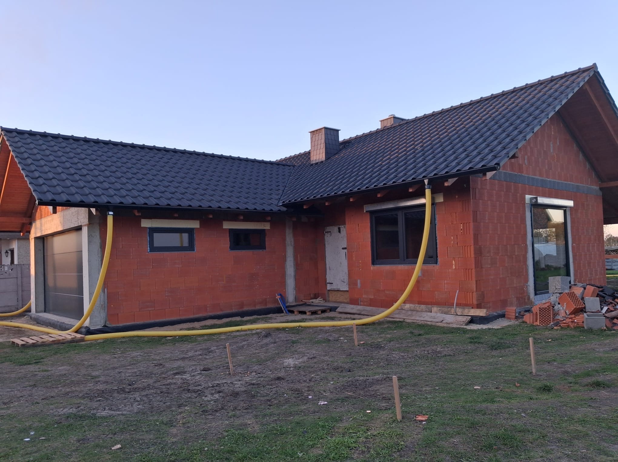 Niedokończony dom z czerwonej cegły z czarnym dachem, widoczne okna i żółte rury wentylacyjne. Częściowo zagospodarowany teren wokół budynku.