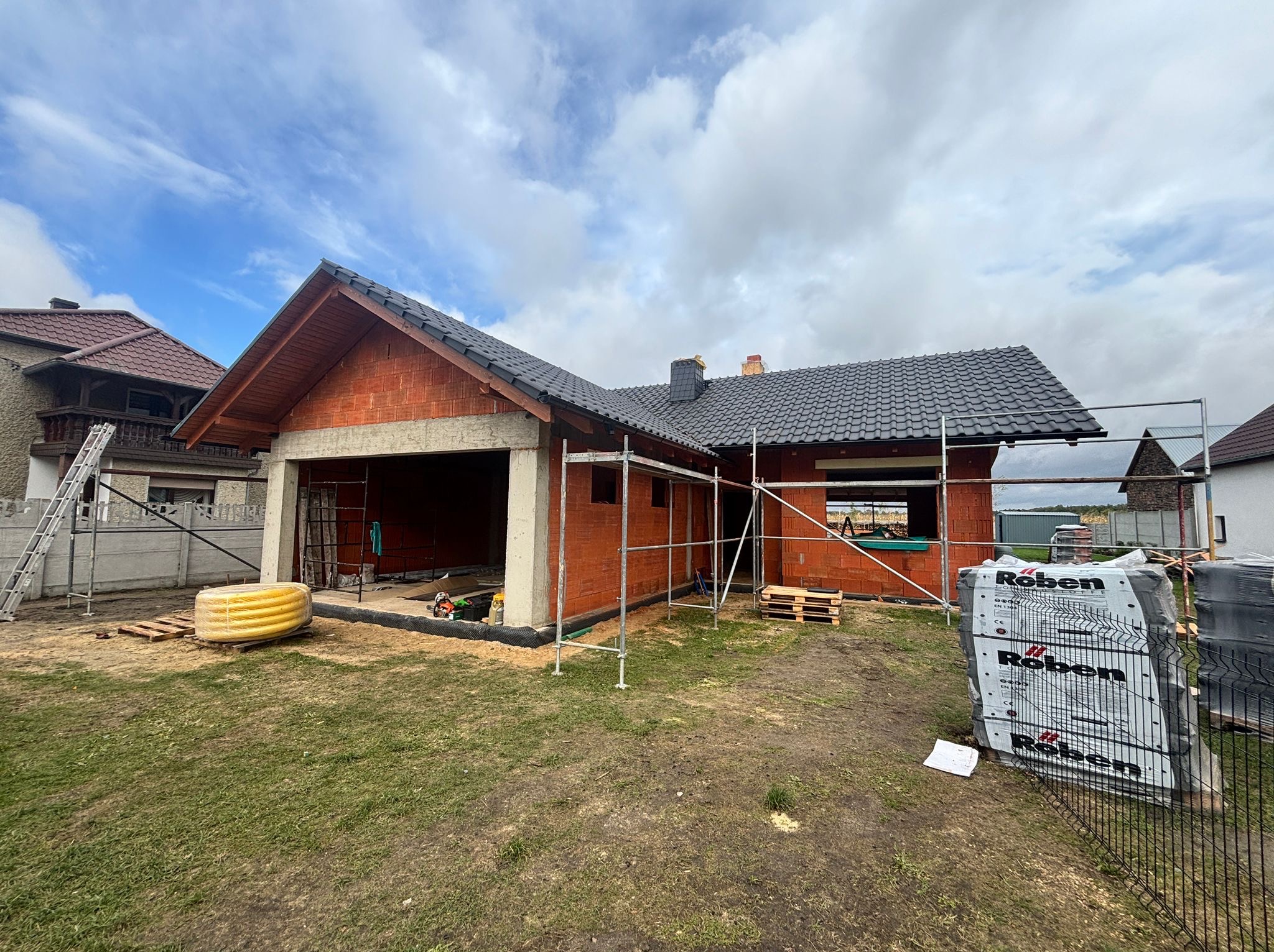 Budowa domu z czerwonej cegły Roben, widoczne rusztowania i materiały budowlane. Dach pokryty czarną dachówką. Niebo pochmurne.