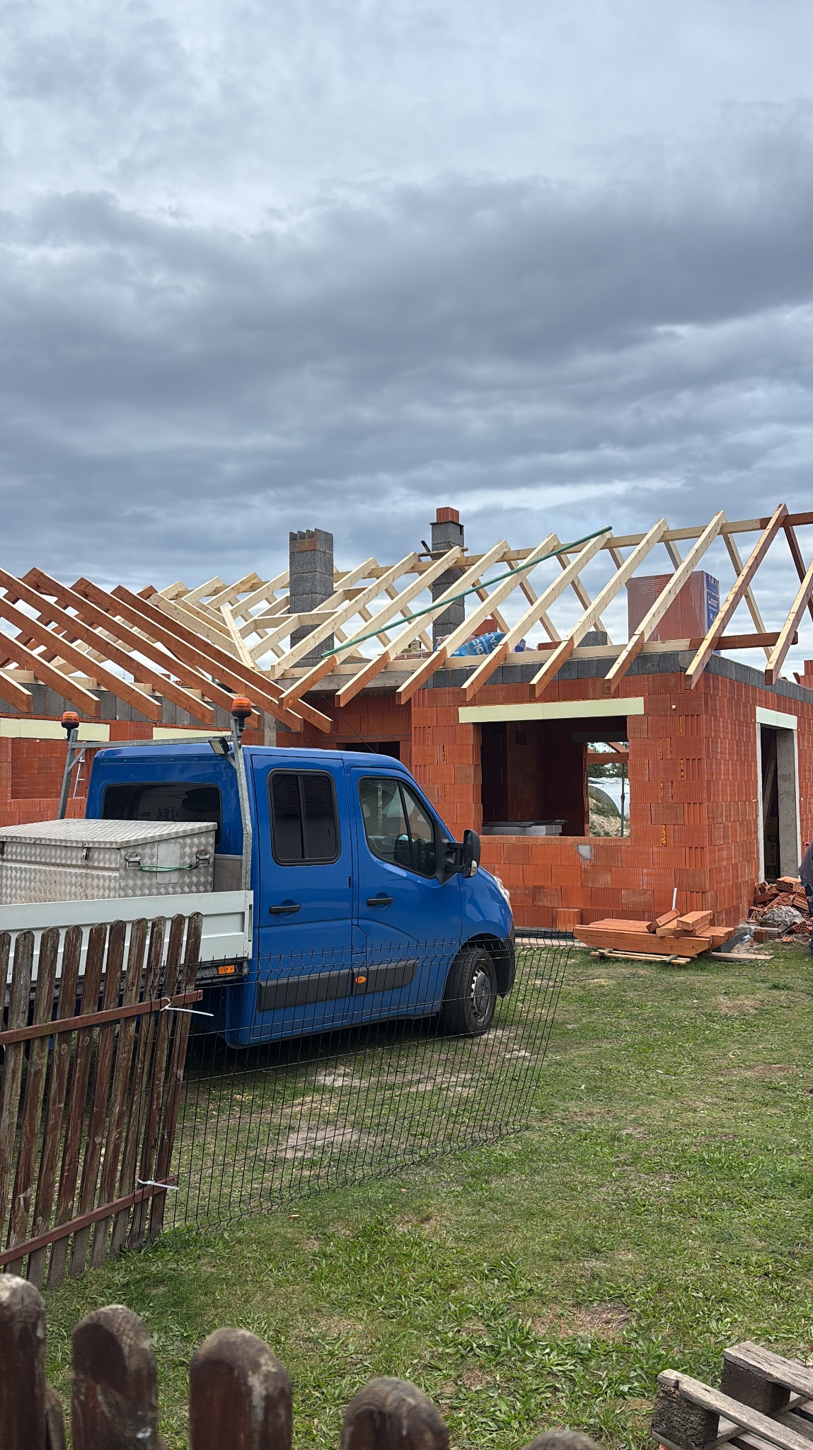 Budowa domu z czerwonej cegły: ściany wzniesione, więźba dachowa w trakcie montażu. Na pierwszym planie niebieski samochód dostawczy z narzędziami.