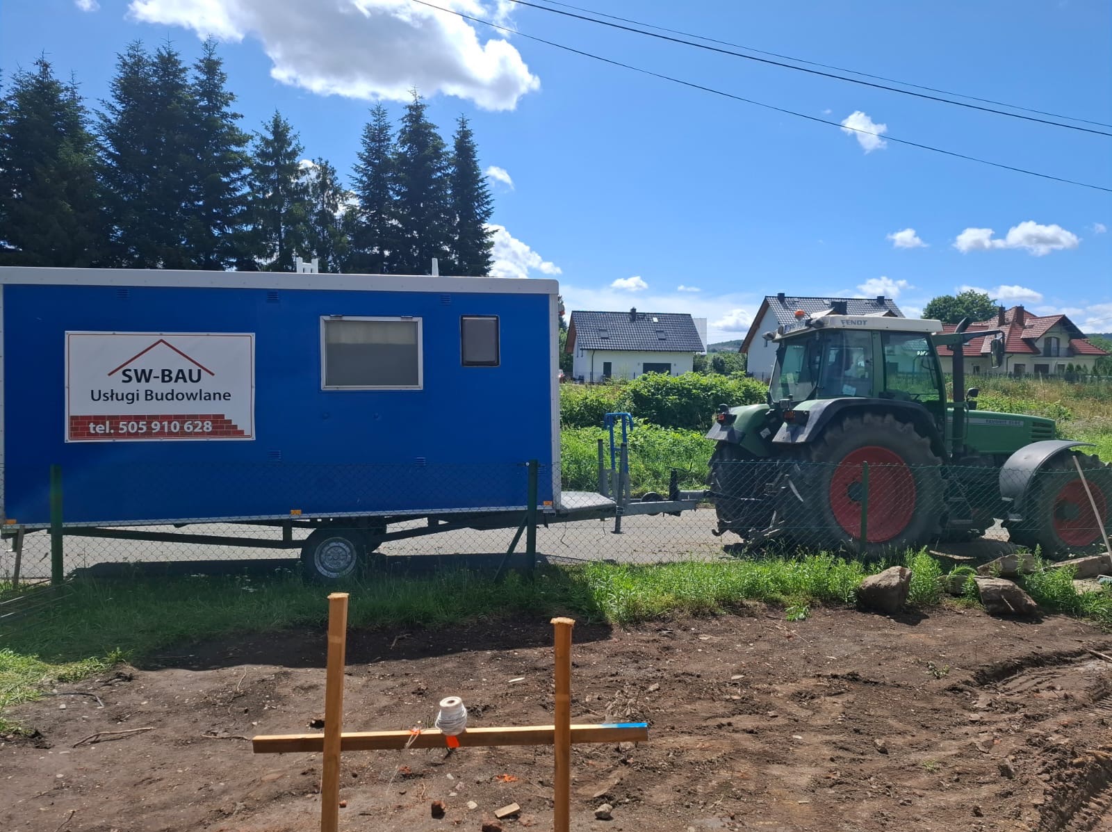 Niebieski kontener biurowy SW-BAU z numerem telefonu na placu budowy, obok zielony traktor Fendt. W tle domy i drzewa pod błękitnym niebem.