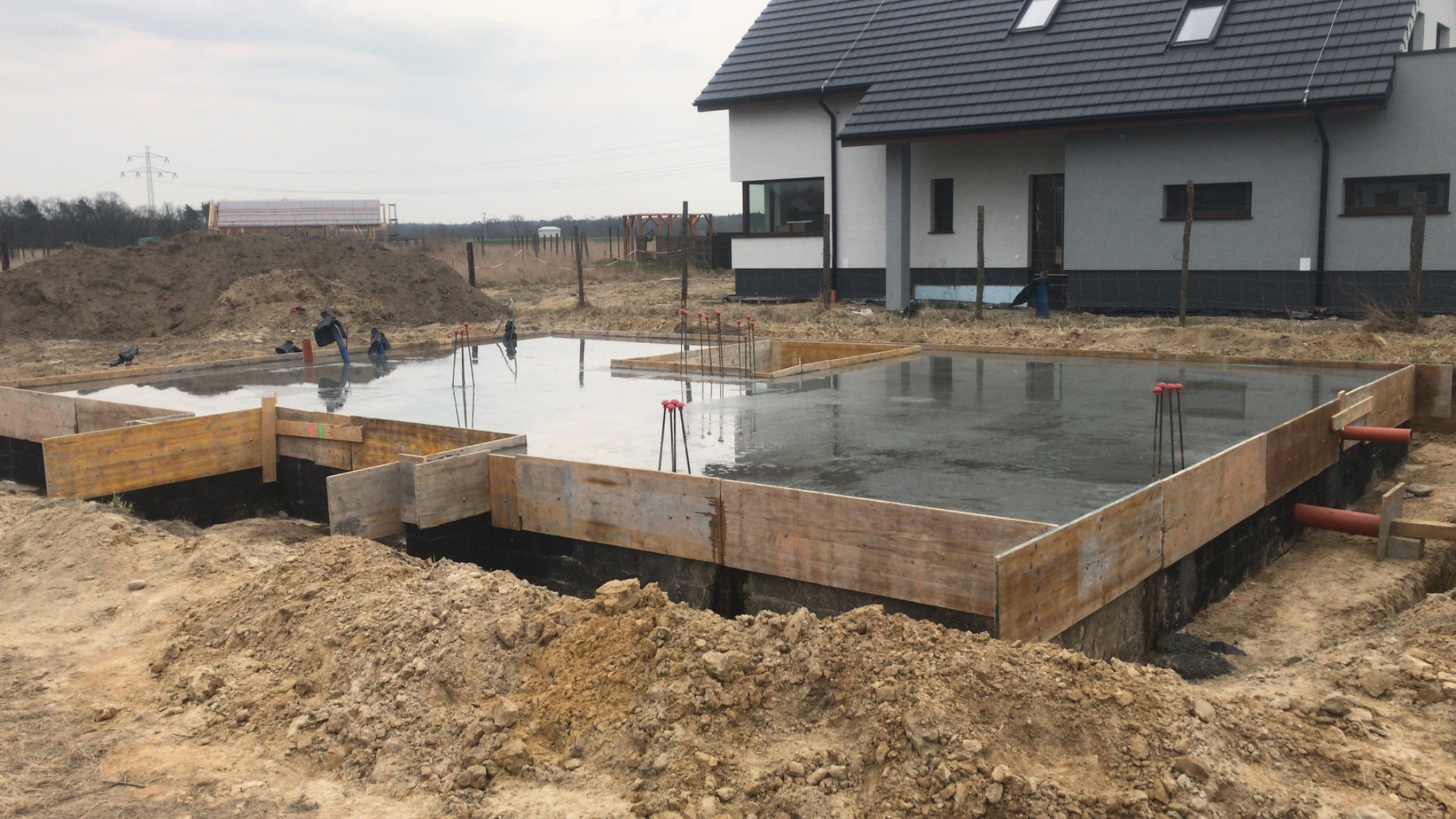 Wylany fundament pod budynek jednorodzinny, widoczne szalunki drewniane, wystające pręty zbrojeniowe i fragment budynku w tle.