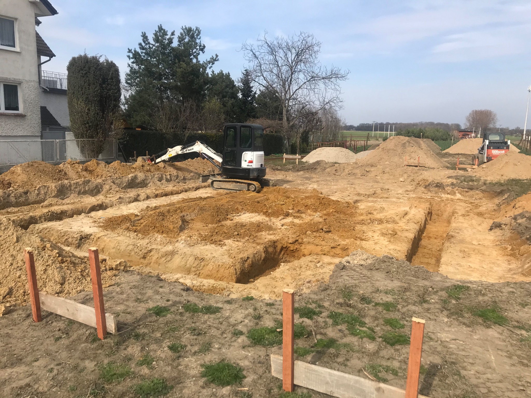 Wykop pod fundamenty domu jednorodzinnego z widocznymi wykopanymi rowami, drewnianym szalunkiem i dwoma minikoparkami Bobcat na placu budowy w słoneczny dzień.