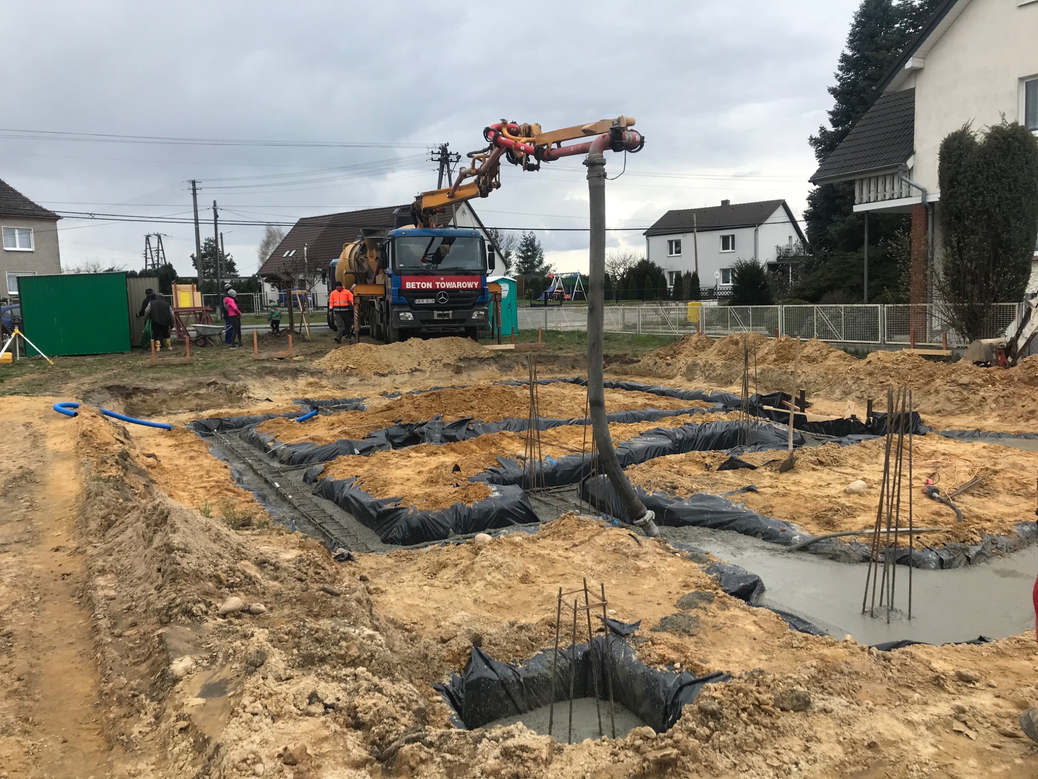 Wylewanie betonu do wykopów fundamentowych z użyciem pompy do betonu, widoczne zbrojenia i izolacja wykopów, w tle zabudowa jednorodzinna i pochmurne niebo.