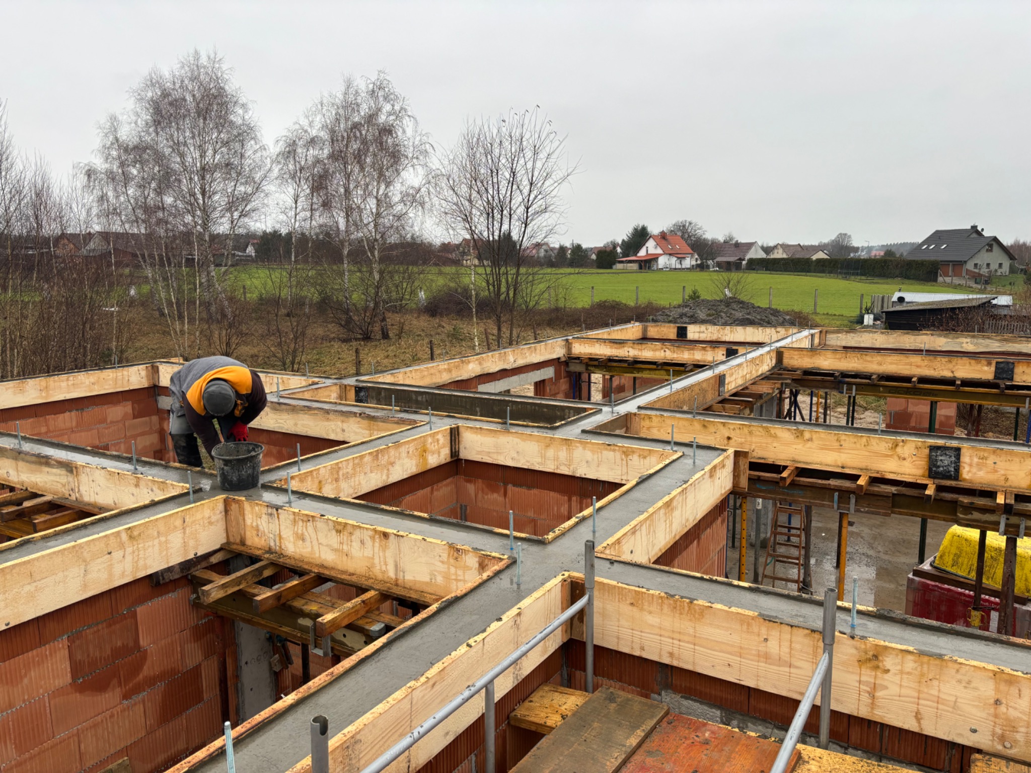 Wypełnianie betonem szalunków fundamentowych pod dom z czerwonej cegły, widok z góry, w tle zabudowa wiejska i nagie drzewa.