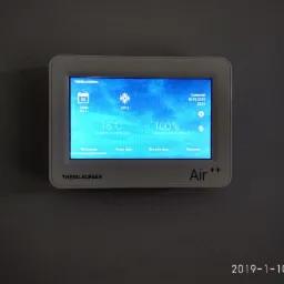 Panel sterowania systemem wentylacji Thesslagreen Air++ zamontowany na szarej ścianie, wyświetlający temperaturę wewnętrzną 16 stopni Celsjusza oraz informację o 100% intensywności wentylacji.