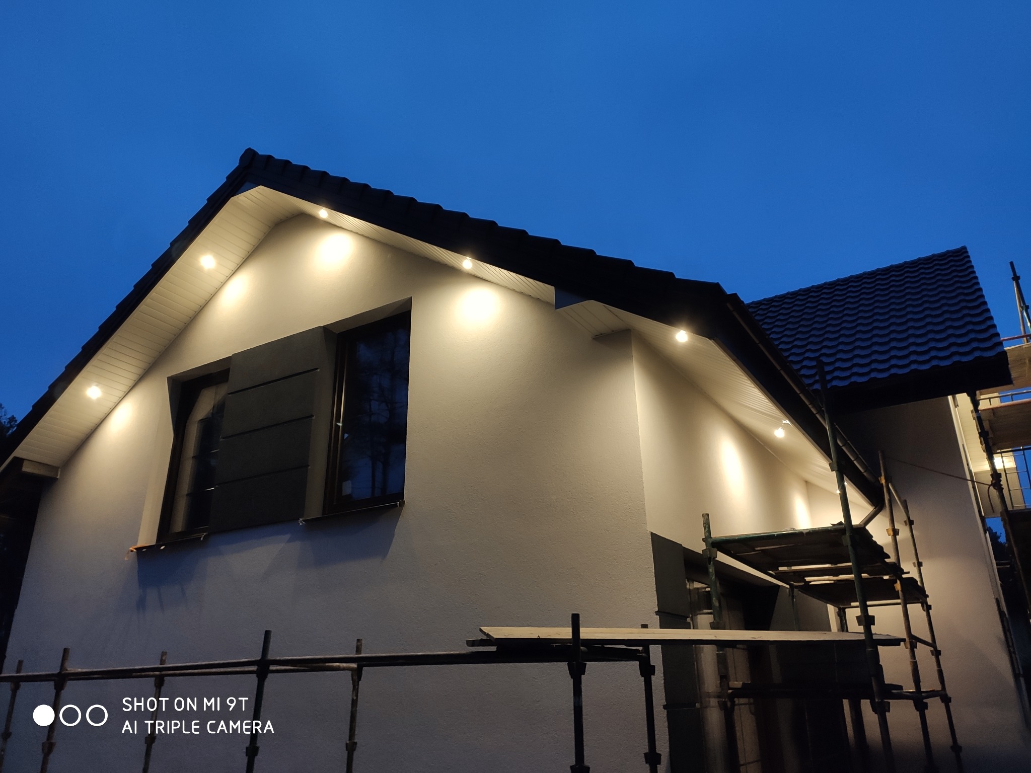 Oświetlony dom jednorodzinny z ciemną dachówką, widoczne rusztowanie, wieczorne oświetlenie elewacji i podbitki dachowej.