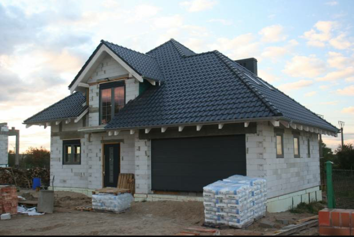 Dom w trakcie budowy z ciemną dachówką, widoczne worki z materiałami budowlanymi i częściowo wykończona elewacja.