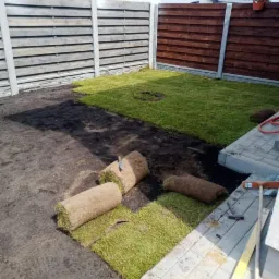 Rozwinięte i zwinięte rolki trawy na ziemi w ogrodzie z częściowo ułożonym trawnikiem, widoczny drewniany płot i narzędzia.
