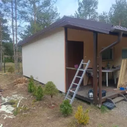 Ocieplona ściana budynku z brązowym dachem, widoczna drabina, tuje i resztki drewna na ziemi. Prace wykończeniowe w ogrodzie.