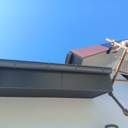 Firma Usługowa Pucuś Adriana Draźba - Szary system rynnowy z bliska, zamontowany na białej elewacji domu. W tle błękitne niebo i pracownik na rusztowaniu malujący fragment dachu na ciemnoszaro.