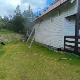 Firma Usługowa Pucuś Adriana Draźba - Czyszczenie elewacji drewnianego domu myjką ciśnieniową z drabiny. Widoczna piana, trawnik i niebo. Osoba w trakcie pracy.