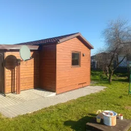 Drewniany domek narzędziowy z brązowym dachem i anteną satelitarną, otoczony zielonym trawnikiem i drzewem. Widoczne farby sugerują świeże pomalowanie.
