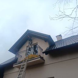 Firma Usługowa Pucuś Adriana Draźba - Praca na wysokości: mężczyzna na drabinie przy balkonie, prawdopodobnie naprawia lub czyści rynnę w domu z beżową elewacją i ciemnym dachem. Widoczne gałęzie drzew.