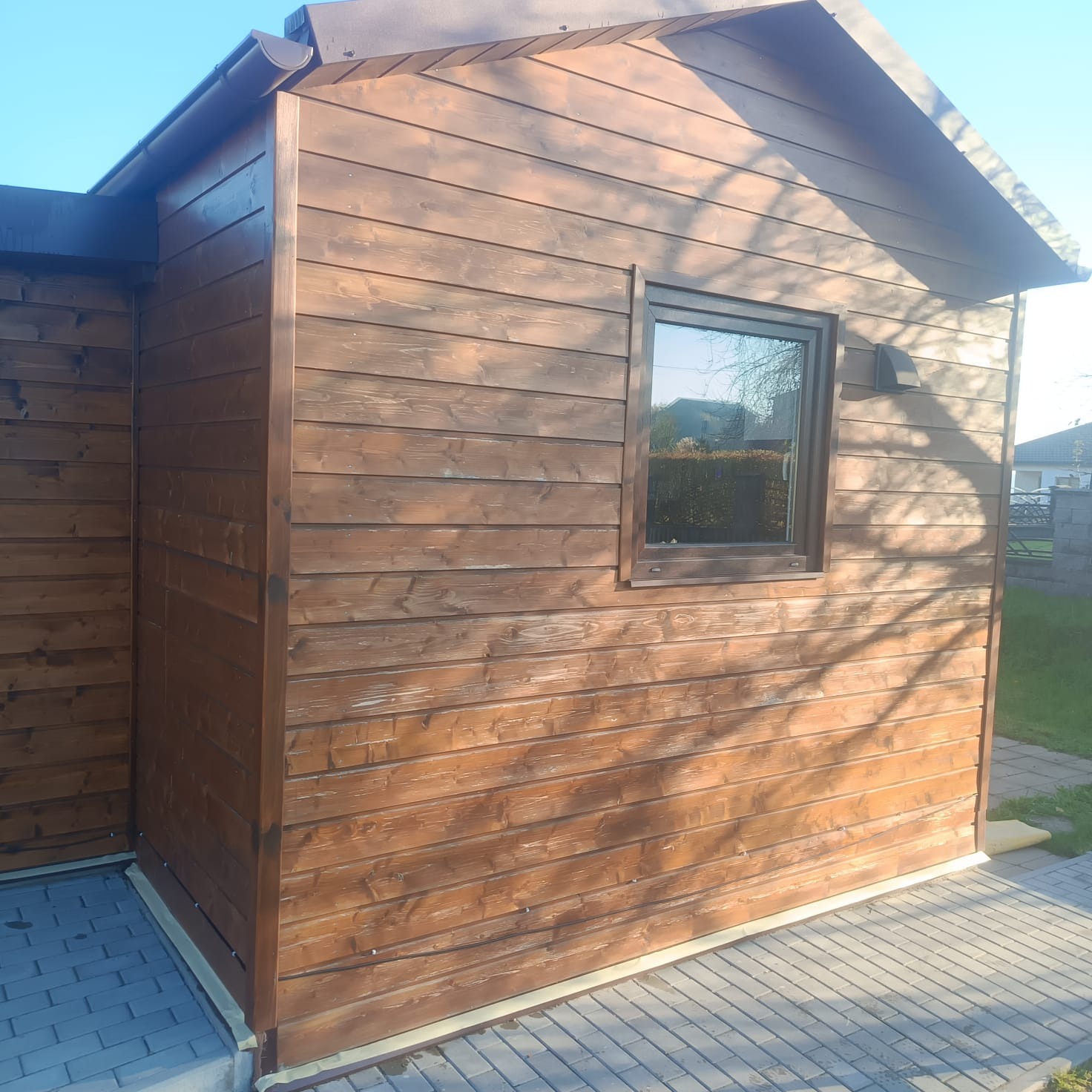 Drewniana altana z oknem, świeżo po malowaniu, z widoczną fakturą drewna i refleksami światła. Detal wykończenia z taśmą malarską przy fundamencie.