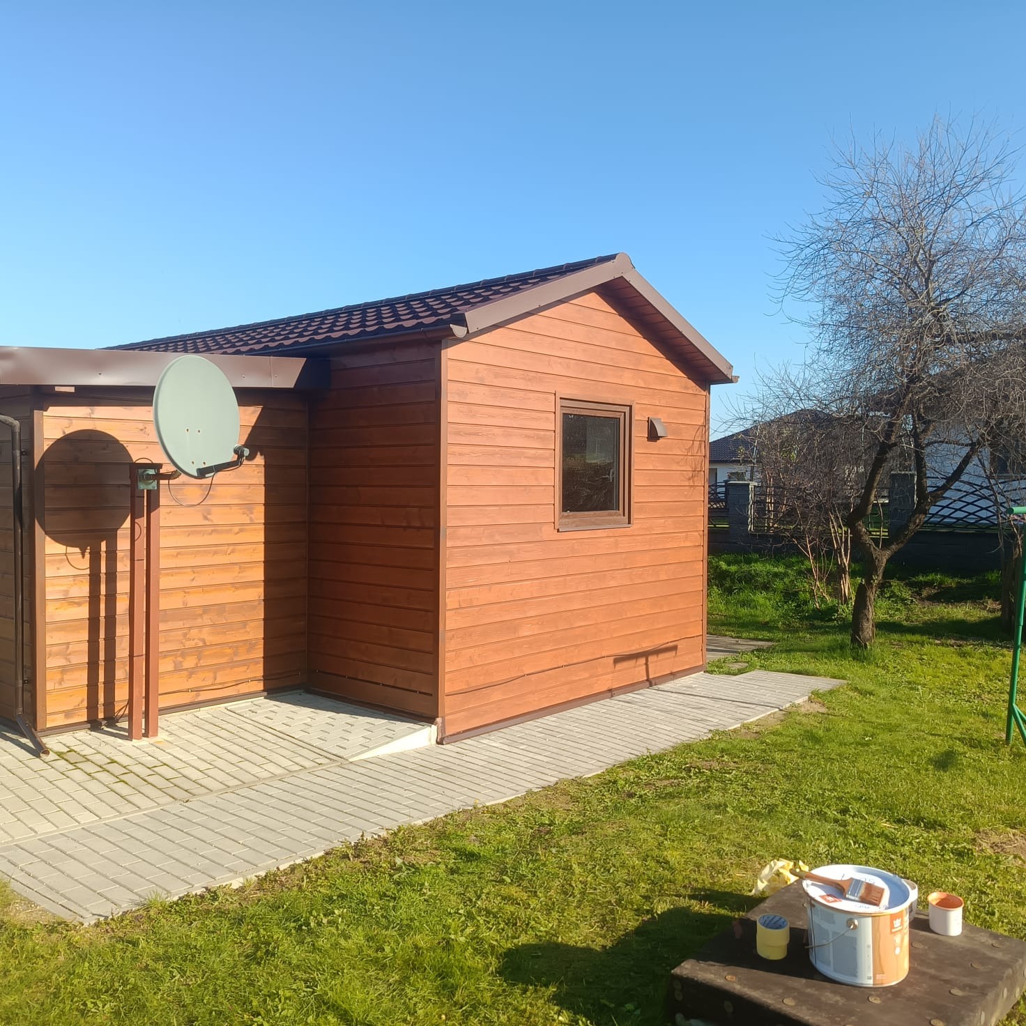 Drewniany domek narzędziowy z brązowym dachem i anteną satelitarną, otoczony zielonym trawnikiem i drzewem. Widoczne farby sugerują świeże pomalowanie.