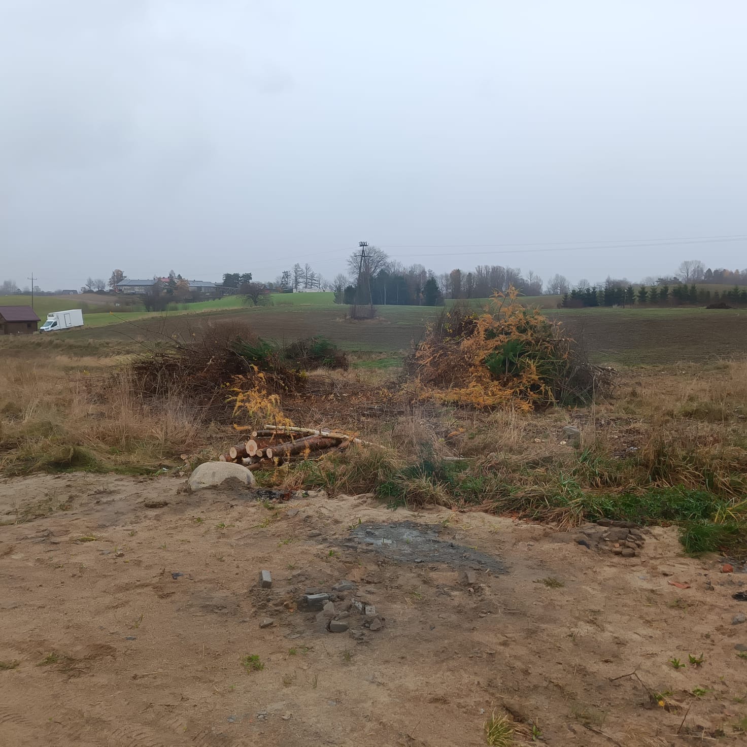 Teren po wycince drzew: stosy gałęzi, pnie drzew leżące obok kamienia, w tle pole i zabudowania w pochmurny dzień. Widoczne ślady po pracy.
