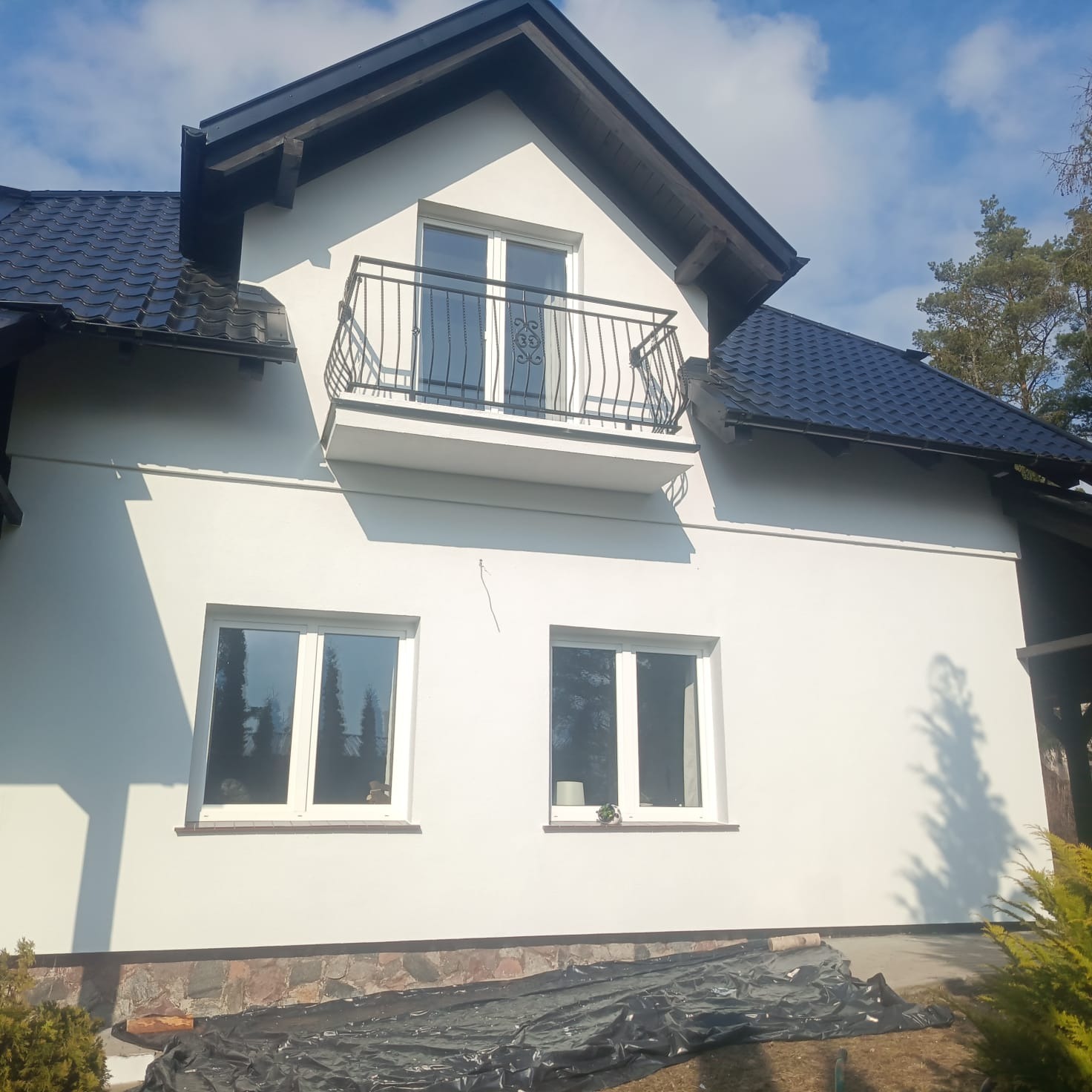 Biała elewacja domu jednorodzinnego z balkonem i ciemnym dachem, po renowacji. Widoczne okna i fragment ogrodu z folią ochronną.