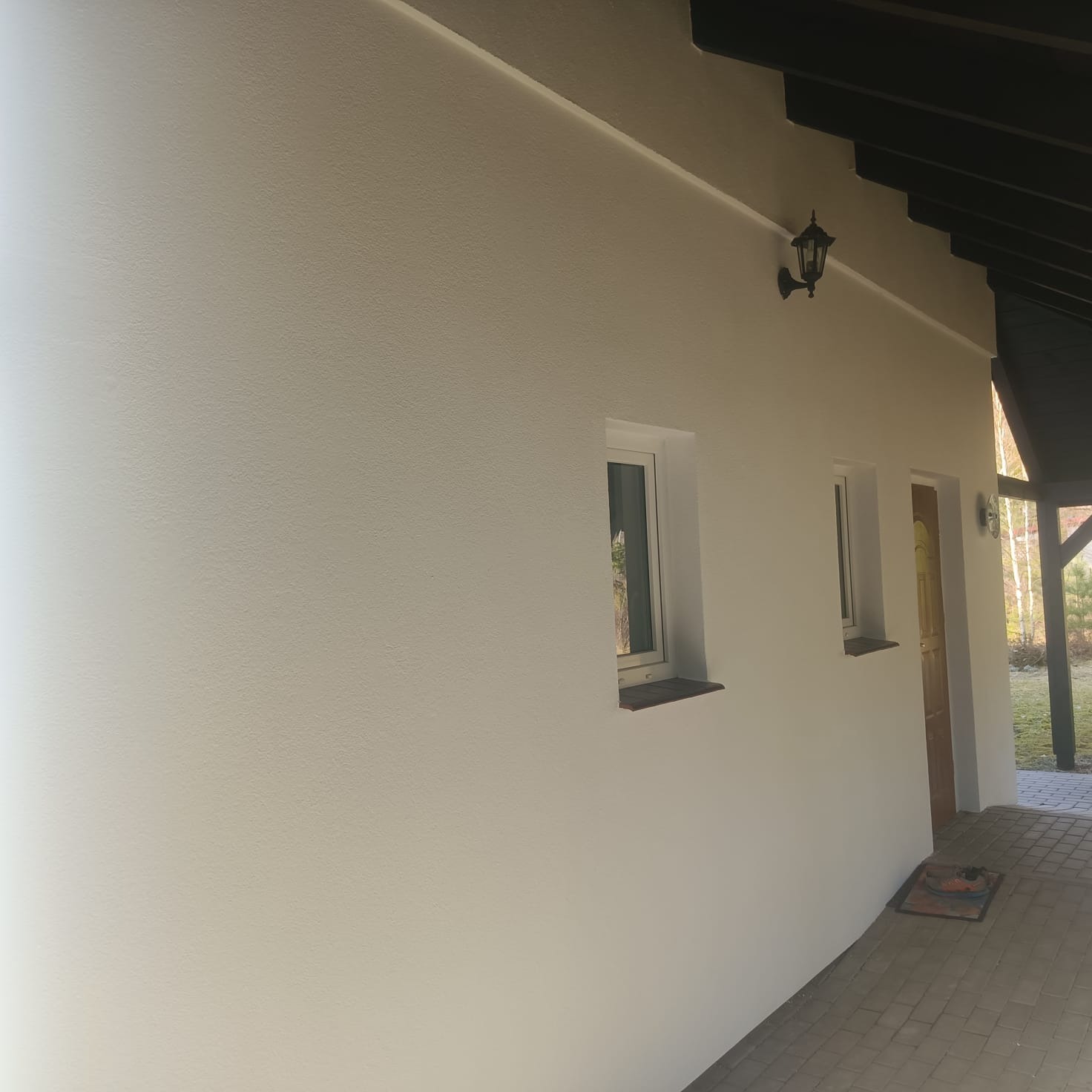 Boczny widok na świeżo pomalowaną, jasną elewację domu z dwoma oknami i drzwiami wejściowymi. Widoczna lampa zewnętrzna i fragment chodnika z kostki.
