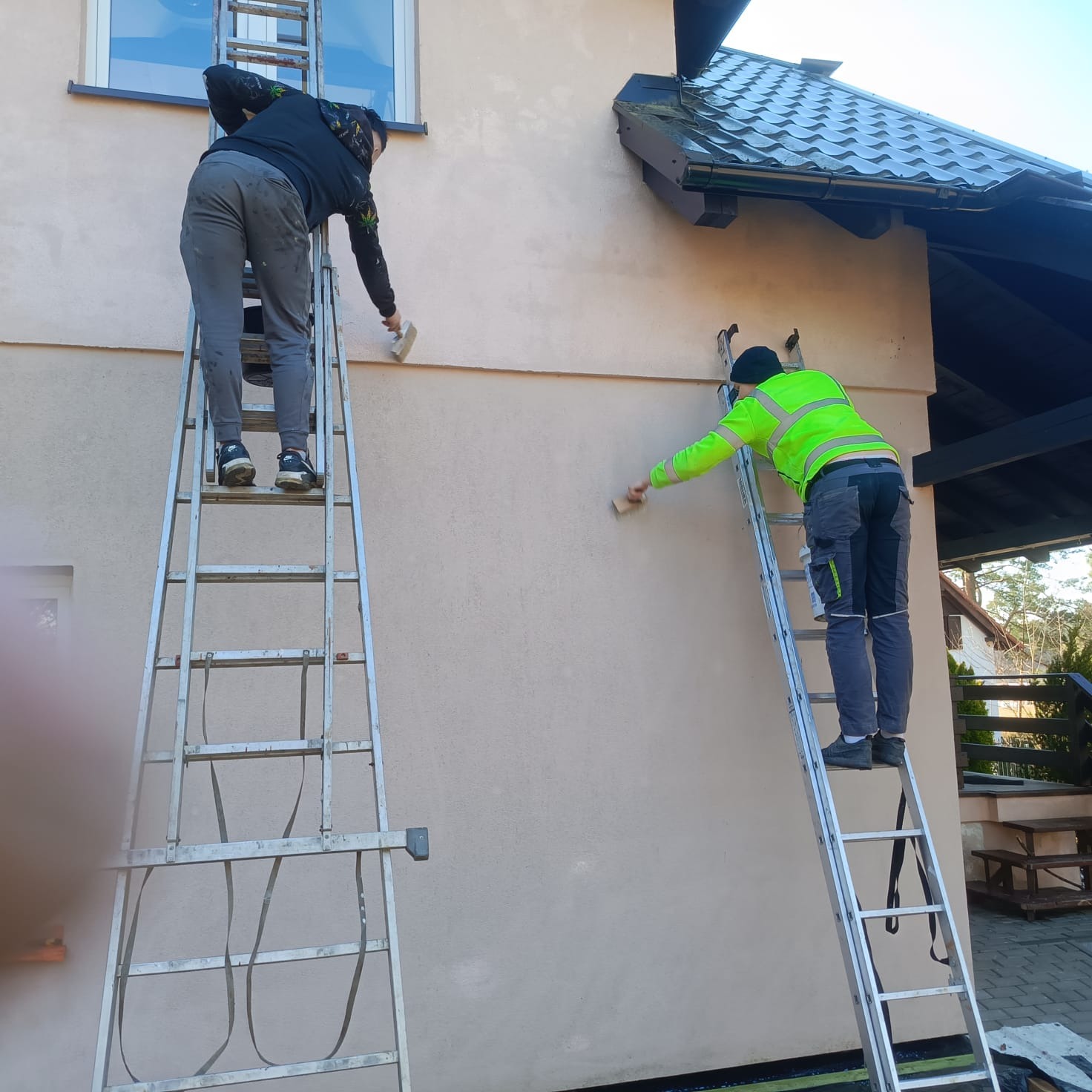 Dwóch pracowników na drabinach przygotowuje elewację domu do malowania. Jeden w neonowej kamizelce, drugi w ciemnej bluzie. Widoczne narzędzia i fragment dachu.