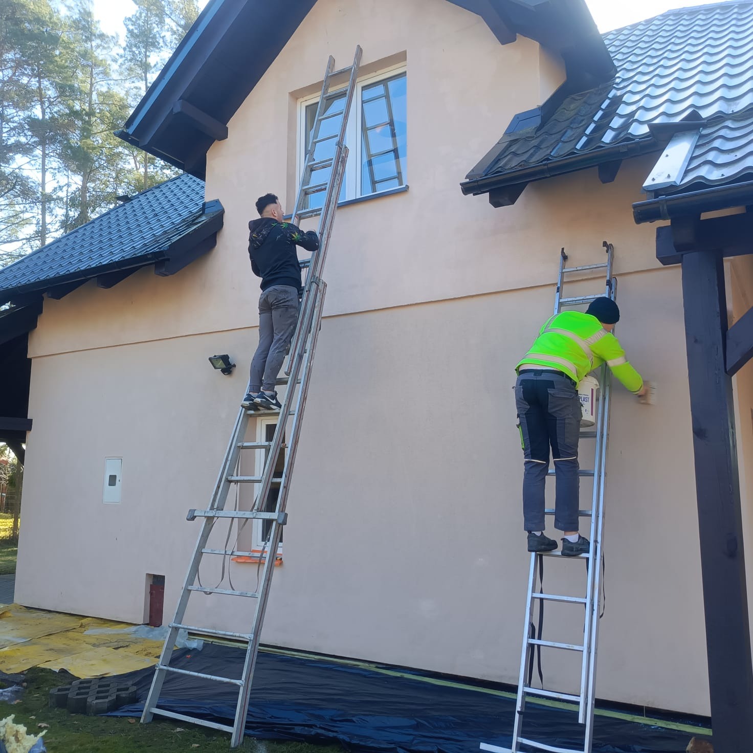 Dwóch mężczyzn na drabinach maluje elewację domu w słoneczny dzień. Jeden maluje okolice okna, drugi górną część ściany. Widoczne narzędzia i zabezpieczenia.