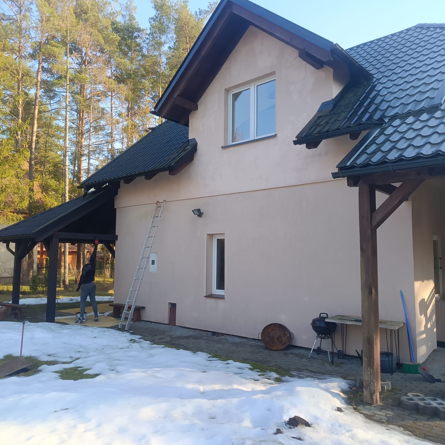 Jasna elewacja domu jednorodzinnego z ciemnym dachem, drabina oparta o ścianę, osoba malująca fragment ściany. Wokół resztki śniegu, drzewa w tle.