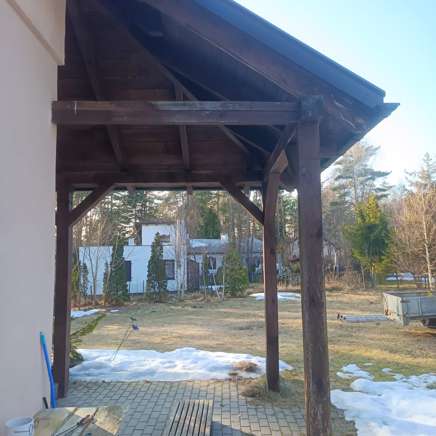 Drewniana altana w Kościerzynie z widokiem na ogród i resztki śniegu. Konstrukcja z ciemnego drewna, częściowo zadaszona, na tle domu i drzew.